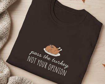 Könnte beinhalten: Ein dunkelbraunes T-Shirt mit dem weißen Text "pass the turkey NOT YOUR OPINION". Das Shirt zeigt einen Cartoon-Truthahn mit Sonnenbrille auf einem weißen Teller. Das Shirt liegt auf einer Holzoberfläche mit Herbstblättern.