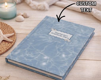 Könnte beinhalten: Ein blaues Dankbarkeitstagebuch mit einem wasserähnlichen Muster. Das Tagebuch trägt den Text "Stephanie's Gratitude Diary" auf einem Etikett. Eine brennende Kerze, Muscheln und ein Seestern befinden sich im Hintergrund.