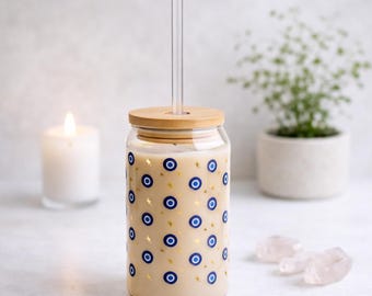Könnte beinhalten: Ein durchsichtiger Glasbecher mit Holzdeckel und klarem Strohhalm. Der Becher ist mit blauen Evil-Eye-Symbolen und goldenen Sternen verziert. Das Glas enthält ein helles Getränk. Eine brennende Kerze und eine Topfpflanze befinden sich im Hintergrund.