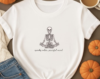 Pode incluir: T-shirt branco com uma ilustração a preto e cinzento de um esqueleto em posição de ioga. O texto "spooky vibes, peaceful mind" está por baixo. A t-shirt é rodeada por abóboras e uma chávena de café.