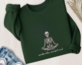 May include: Dark green sweatshirt with a white skeleton illustration in a yoga pose. The text "spooky vibes, peaceful mind" is printed below. The sweatshirt has a crew neck and long sleeves.