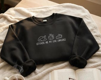 May include: Black sweatshirt with white graphic of a turkey, pumpkin, and pie slice, with the text "Leftovers are my love language." The sweatshirt is on a cream-coloured blanket, with a cup of coffee and an open book with glasses.
