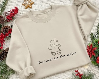 Könnte beinhalten: Beiges Sweatshirt mit Rundhalsausschnitt und langen Ärmeln. Auf der Vorderseite befindet sich eine schwarze Strichzeichnung eines Lebkuchenmanns mit Schleife. Darunter steht der Text "Too sweet for this season".
