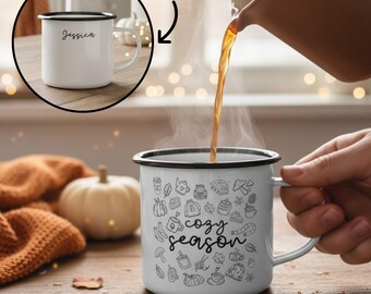Könnte beinhalten: Weißer Emaillebecher mit schwarzem Rand und Henkel, mit den Worten "cozy season" und herbstlichen Kritzeleien. Eine Person gießt ein heißes Getränk in den Becher. Der Becher im Inset trägt den Namen "Jessica".