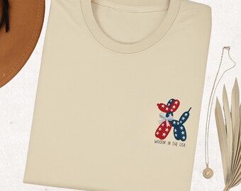 May include: A cream-coloured t-shirt with a patriotic balloon dog design in red, white, and blue stars. The text "Woofin' in the USA" is printed below the graphic. The t-shirt is folded.