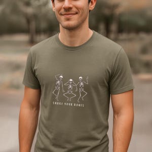 May include: An olive green t-shirt with a white graphic of three dancing skeletons. The text "SHAKE YOUR BONES" is printed below the skeletons. The shirt has a crew neck and short sleeves, a casual style.