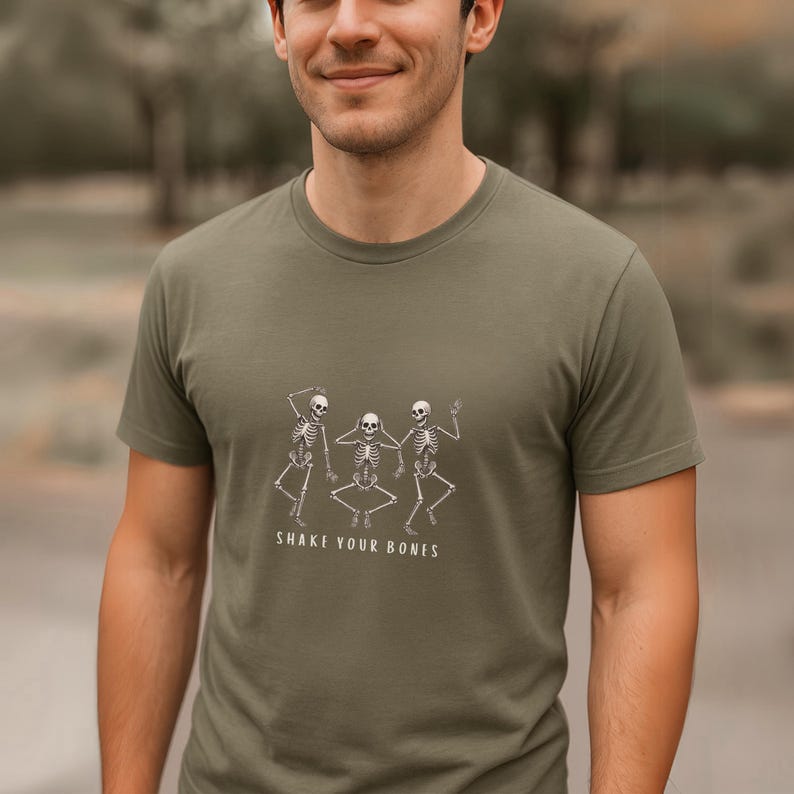May include: An olive green t-shirt with a white graphic of three dancing skeletons. The text "SHAKE YOUR BONES" is printed below the skeletons. The shirt has a crew neck and short sleeves, a casual style.