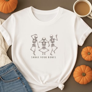 May include: White t-shirt with a graphic of three dancing skeletons. The skeletons are in a line, with one covering their eyes, one covering their ears, and one with their arms raised. The text "SHAKE YOUR BONES" is below the skeletons.