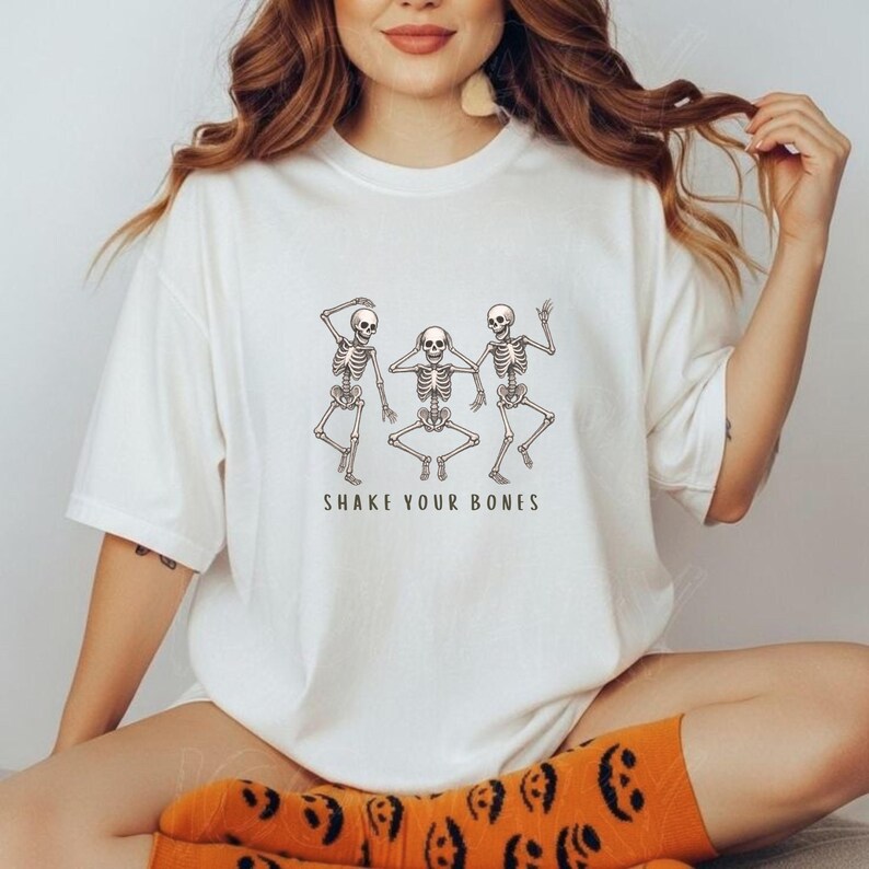 May include: White t-shirt with a graphic of three dancing skeletons. The skeletons are in different poses, with the text "SHAKE YOUR BONES" underneath. The shirt has a relaxed, oversized fit.