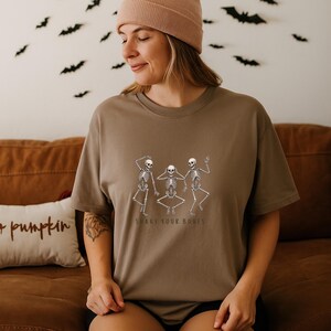 May include: A brown t-shirt with a graphic of three dancing skeletons and the text "SHAKE YOUR BONES". The shirt is worn by a person wearing a beige beanie. Bats are visible in the background.