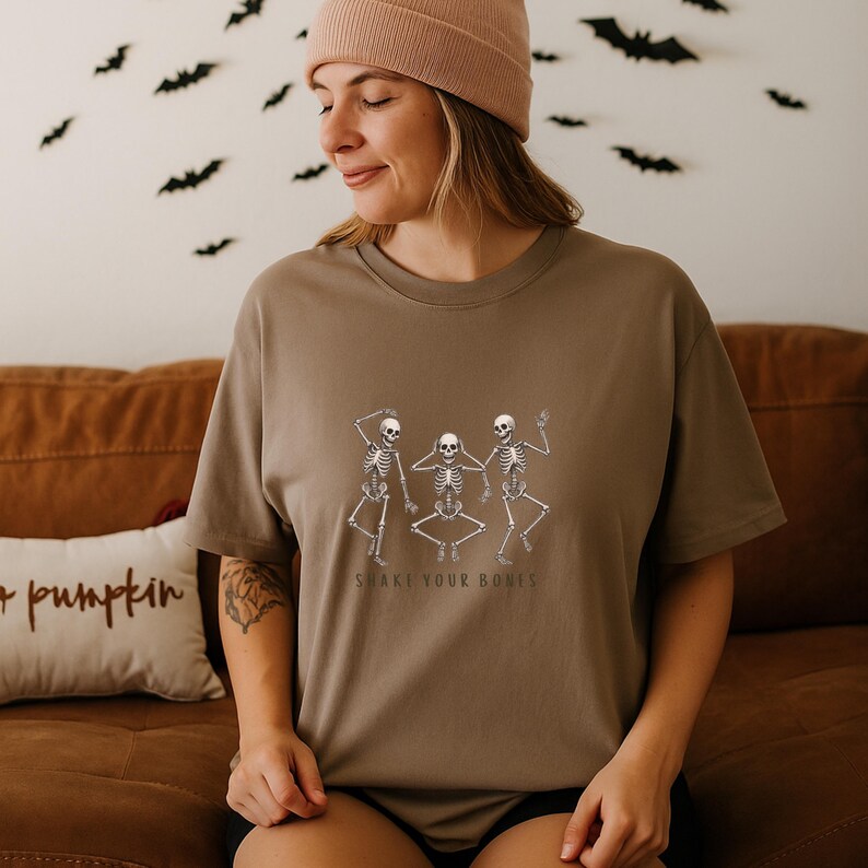 May include: A brown t-shirt with a graphic of three dancing skeletons and the text "SHAKE YOUR BONES". The shirt is worn by a person wearing a beige beanie. Bats are visible in the background.