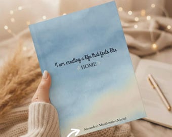 Könnte beinhalten: Ein rechteckiges Journal mit einem blau-weißen Aquarell-Verlauf auf dem Einband. Auf der Vorderseite steht der schwarze Text: I am creating a life that feels like HOME, sowie der Zusatztext: Alexandra's Manifestation Journal.