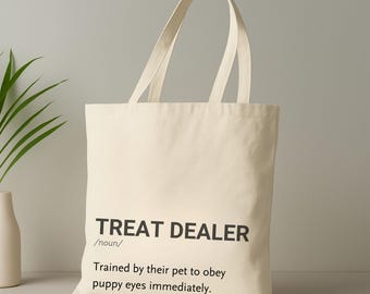 Könnte beinhalten: Cremefarbene Canvas-Tragetasche mit schwarzem Text: "TREAT DEALER /noun/ Trained by their pet to obey puppy eyes immediately." Die Tasche hat zwei lange Henkel und ist zum Tragen von Gegenständen geeignet.