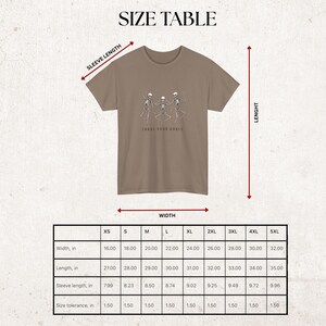 May include: A tan t-shirt featuring a graphic of three dancing skeletons and the text "Shake Your Bones." The image includes a size table with measurements in inches for width, length, and sleeve length.