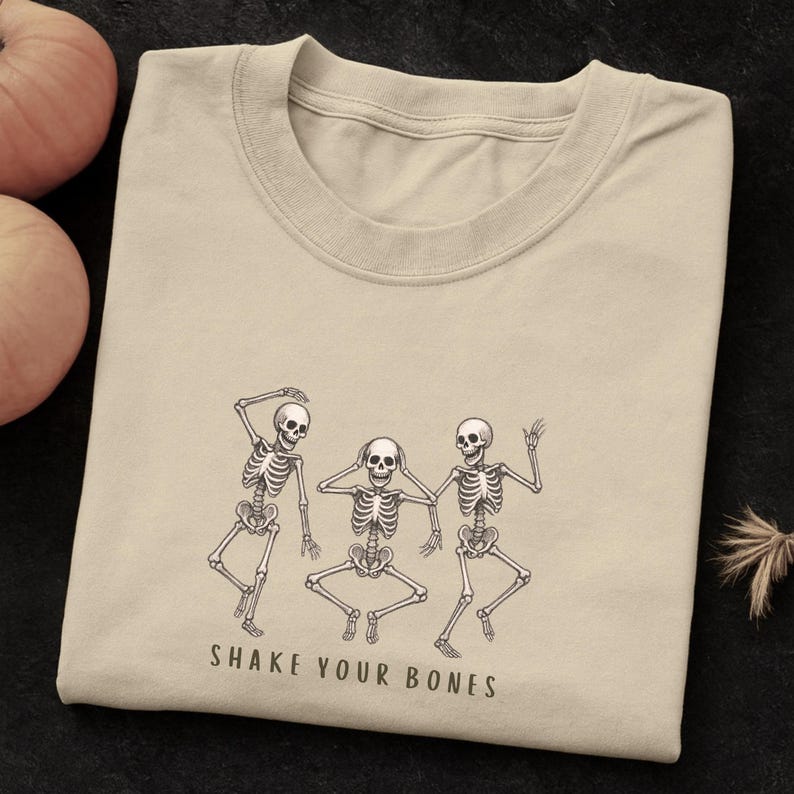 May include: A beige t-shirt with a black and white illustration of three dancing skeletons. The text "SHAKE YOUR BONES" is printed below the skeletons. The shirt is folded and has a crew neck.