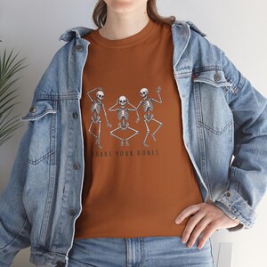 May include: A rust-coloured t-shirt with a graphic of three dancing skeletons. The skeletons are in various poses, with the text "SHAKE YOUR BONES" printed below them in black. The shirt is made of a soft material.