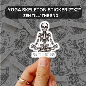 Op de afbeelding: Een witte sticker met een mediterend skelet in een yogahouding. De sticker heeft de tekst "YOGA SKELETON STICKER 2"X2"" en "ZEN TILL' THE END". De sticker wordt vastgehouden door een hand met een gouden ring.