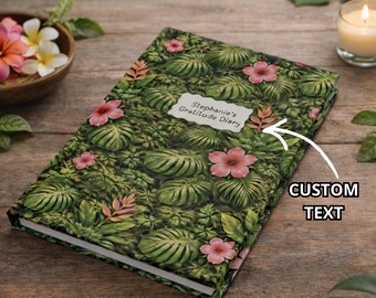 Könnte beinhalten: Ein Notizbuch mit einem Muster aus tropischen Blättern und rosa Blüten. Auf dem Einband steht "Stephanie's Gratitude Diary" auf einem weißen Etikett. Eine brennende Kerze und eine Schale mit Blumen befinden sich im Hintergrund.