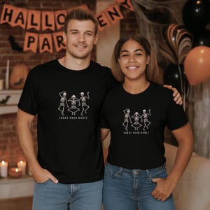 May include: Black t-shirts featuring a dancing skeleton design and the words "Shake Your Bones." The shirts are worn by two people, set against a Halloween backdrop with balloons and a banner.