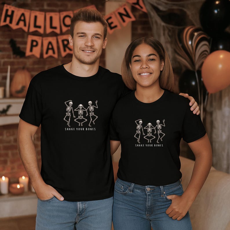 May include: Black t-shirts featuring a dancing skeleton design and the words "Shake Your Bones." The shirts are worn by two people, set against a Halloween backdrop with balloons and a banner.