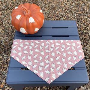 May include: A triangular pet bandana in a dusty rose color featuring a repeating pattern of small white ghosts, displayed alongside a round orange pumpkin decorated with hand-painted white ghost figures on its surface.