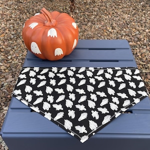 May include: An orange pumpkin with white ghost designs sits next to a black and white ghost-patterned bandana. The bandana is triangular and has a white stitched edge. The items are on a blue surface.