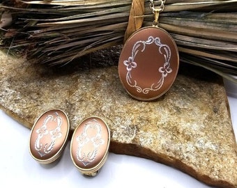 May include: A set of vintage jewellery featuring a gold chain necklace with a brown oval pendant and matching earrings. The pendant and earrings have a white floral design.