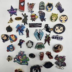 May include: A collection of various metal enamel pins featuring comic book characters, superhero logos, and pop culture icons. The assortment includes figures like Spider-Man, Batman, Iron Man, and Hulk, along with text-based pins like Marvel Collector.