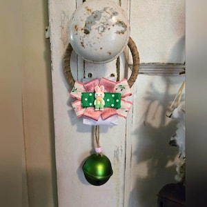 Puede incluir: Adorno de puerta con temática de Pascua con un pomo de puerta blanco desgastado y una corona decorativa. La corona incluye cintas rosas y blancas, una cinta verde con lunares blancos y una pequeña figura de conejito. Una campana verde cuelga debajo.