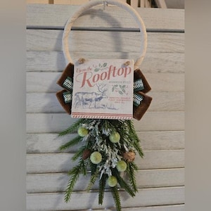 Puede incluir: Una corona navideña con un anillo de cuerda color crema. Presenta un letrero que dice "Upon the Rooftop" con una ilustración de un reno. Tela marrón y detalles verdes rodean el letrero, con vegetación artificial, piñas y adornos de bolas verdes.