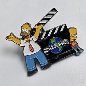 May include: A colorful enamel pin featuring Homer Simpson with arms outstretched, a film clapperboard, and Bart Simpson. The clapperboard has the Universal Studios logo. The pin is a collectible item.