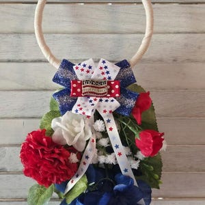 Op de afbeelding: Een patriottische krans met een crèmekleurige touwring, versierd met een rood, wit en blauwe strik. De strik heeft een Amerikaanse vlagspeld en sterrenpatroon linten. De krans bevat rode, witte en blauwe kunstbloemen.