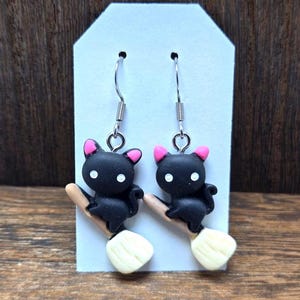 Puede incluir: Un par de pendientes temáticos de Halloween con figuras de gatos negros con orejas rosas y ojos blancos, posados sobre escobas en miniatura. Los pendientes tienen ganchos plateados y están colocados sobre una etiqueta blanca, perfectos para la temporada.