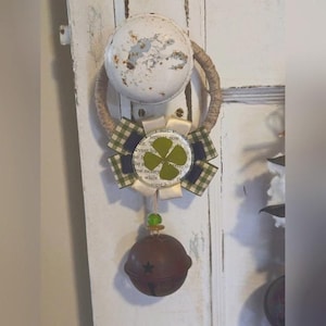 Puede incluir: Un trébol de cuatro hojas verde con una cinta a cuadros blanca y azul cuelga de un pomo de puerta rústico con una campana marrón unida a la parte inferior.