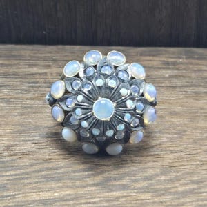 May include: A silver-toned ring with a floral design, featuring multiple oval and round opals. The opals are a mix of white and pale blue, set against a dark metal base. The ring is on a wooden surface.