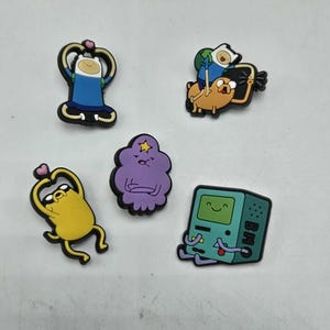 May include: A selection of five cartoon character charms. The charms depict characters in different poses, including Finn, Jake, BMO, and Lumpy Space Princess. The charms are brightly coloured with black outlines, ideal for fans of the animated series.