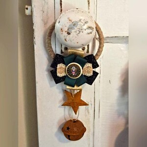 May include: A decorative door knob hanger featuring a white and speckled knob, a bow with a US Army emblem, a star, and a rusty bell. The bow has black, green, and gold accents. The star and bell are made of metal.