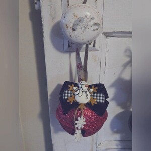 Può includere: Un ornamento a forma di campana di Natale rosso glitterato con un fiocco a scacchi bianco e nero, un pupazzo di neve e fiocchi di neve.