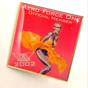 May include: A pink and gold pin featuring a silver robot woman wearing a yellow dress. The pin reads "Aero Force One Official Member" and "2002".