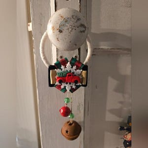 May include: A Christmas ornament featuring a red truck carrying a Christmas tree, set against a snowflake and plaid background. The ornament hangs from a white loop and includes red and green beads, and a small bell.