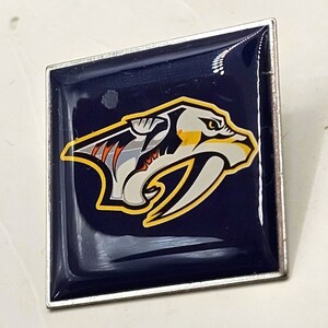 May include: A silver and blue enamel pin featuring the Nashville Predators NHL team logo. The logo is a stylized predator with yellow and orange accents.