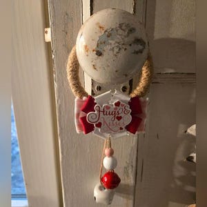 Puede incluir: Un pomo de puerta decorativo con un acento de cuerda y un letrero que dice "Hugs & Kisses". El letrero tiene un lazo rojo y blanco, con una cadena de cuentas y una campana roja colgando debajo. El pomo de la puerta es blanco con un acabado envejecido.
