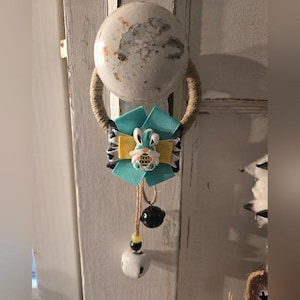 May include: A decorative Easter-themed door knob hanger. The hanger features a teal and yellow ribbon flower with a bunny embellishment. Below the flower are two dangling bells, one black and one white, with a jute rope loop.