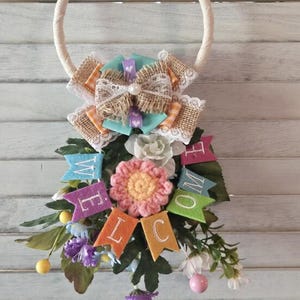 Pode incluir: Uma grinalda decorativa com um anel de corda creme, adornada com um laço de serapilheira, renda e bandeiras de feltro coloridas que dizem "WELCOME". A grinalda apresenta uma flor rosa central, vegetação e pequenos detalhes florais.