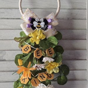 May include: A decorative wreath with a cream-colored rope ring, adorned with a bow, a bee emblem, and faux flowers. The wreath features the words "BEE KIND" spelled out on felt hexagon shapes, with yellow, white, and orange floral accents.