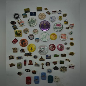 May include: A collection of vintage pins and badges in various shapes and sizes. The pins feature a variety of designs, including text, logos, and illustrations. Colors include red, blue, green, yellow, and silver.