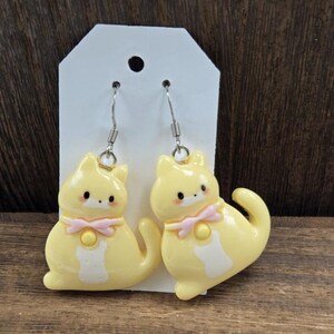 May include: A pair of yellow cat-shaped earrings with pink bows. The earrings are made of polymer clay and have silver hooks.