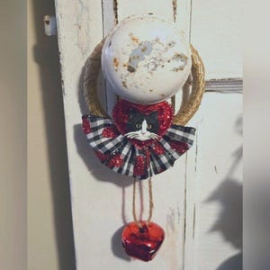 May include: A black and white cat with a red heart and glitter bow hangs from a rustic wreath with a red bell.