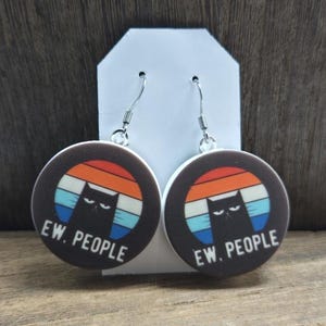 May include: Round earrings with a black cat design, set against a retro sunset of orange, blue, and white stripes. The text "EW. PEOPLE" is printed below the cat. The earrings hang from silver-colored hooks.
