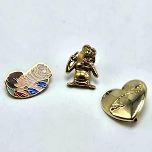 May include: Three vintage enamel pins. One pin is gold-toned and shaped like the character E.T. The second pin is a gold-toned heart with a design. The third pin is an oval shape with a profile of a person and a swirling design.